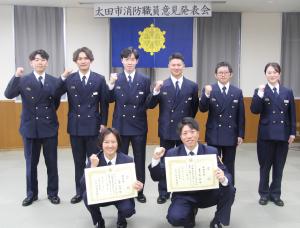 消防職員意見発表会発表者の集合写真。前列右側に藤原副士長、前列左側に荻野消防士がおり、後列に努力賞の６名がいる。全員で右手でガッツポーズをしている写真。